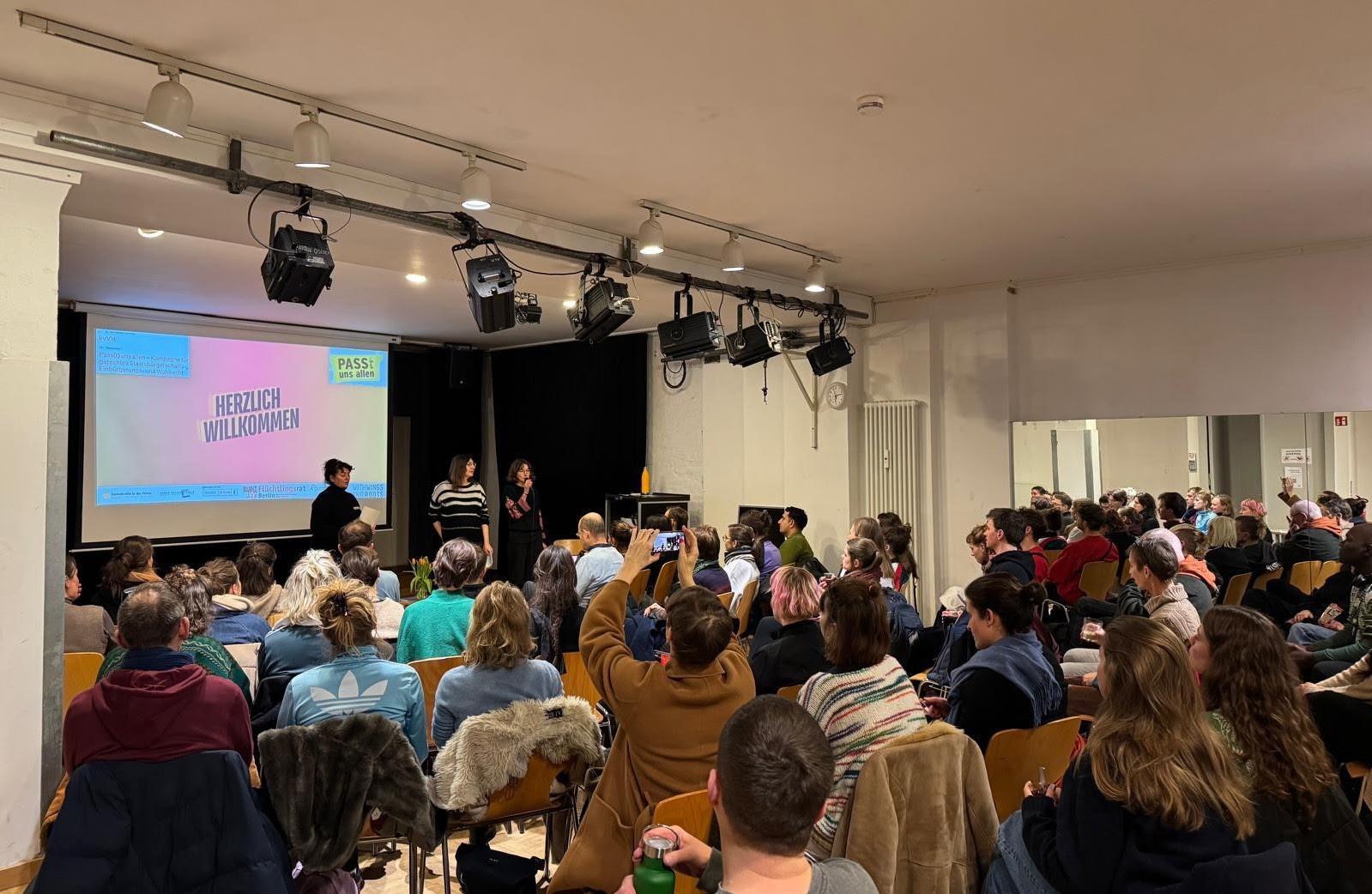 Audience seated in a room watching a presentation on a stage, with a projector screen displaying content, and 3 speakers standing at the front